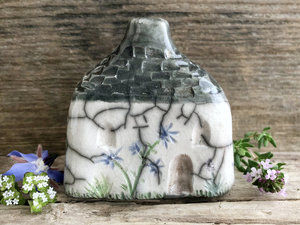Raku ceramic spirit house. The top is grey and carved to look like slates. The rest is a white cottage with windows, a door, and flower and plant carvings. The flowers are a mix of violet, blue, and yellow, and the leaves and stems are green.