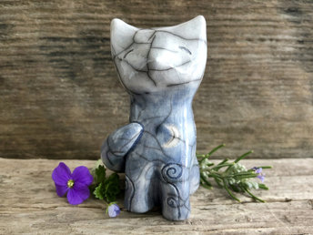 Raku ceramic sitting fox glazed in a bottom-to-top gradient of grey-blue to almost white. It has a kind, gentle, upturned face, and its tail is curled around its back. It has a white crescent moon carving on its chest and spiral designs on its legs and back.