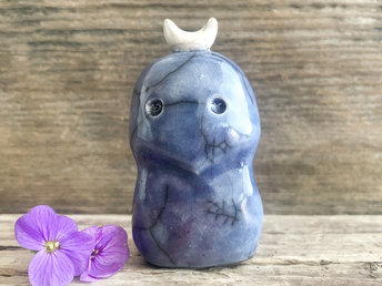 A kodama nature and forest spirit kami ceramic raku sculpture glazed in blue with hints of soft red. It has a gentle face and a white crescent moon on its head.