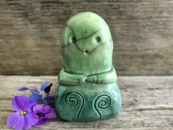 A sweet kodama nature spirit ceramic guardian statue glazed in shades of green and with a lovely, gentle face and little arms. it has cletic spiral designs carved on its front.