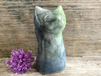 Raku ceramic earth and water spirit kitsune sitting fox sculpture glazed in green, blue, and grey. It is sitting up with its tail curled up its back, and it has a gentle, kind face. 