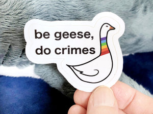 a sticker with a white background and the words "be geese, do crimes". next to the words on the right is a drawing of a slightly vexed looking goose. it has an orange beak and a rainbow neck.