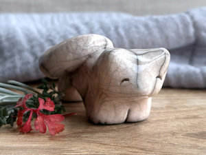 Raku ceramic kitsune fox sculpture glazed in a very pale peach colour. It has a kind, gentle face, and its tail is curled over its back on its right.