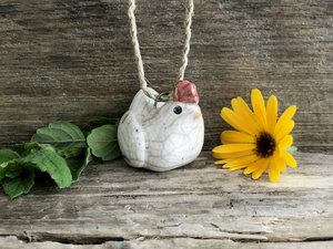 Raku ceramic chicken pendant on eco flax cord. She is glazed in white with an orange beak and a bordeaux-red chicken thingy on her head.