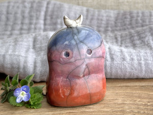 A kodama nature and forest spirit kodama kami ceramic raku sculpture glazed in a bottom-to-top gradient of red-orange to mauve to blue. It has a gentle (if slightly worried) face, and a white crescent moon on its head.