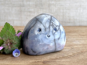 small cloud raku ceramic sculpture glazed in a bottom-to-top gradient of violet to blue to pale blue, almost white at the top. it has an adorable, happy face and is shaped vaguely like a squat raindrop. there are tiny dark grey spiderweb patterns in the glaze, in addition to the normal raku crackles, from where raindrops fell on it.