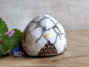 Raku ceramic little owl or owlet kami nature spirit statue glazed in white and brown with gentle, smiling eyes and a little golden yellow beak. There are tiny dark grey spiderweb patterns in the glaze, in addition to the normal raku crackles, from where raindrops fell on it.