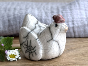 a white raku ceramic chicken with grey eyes, a pale orange beak, and a bordeaux red comb on the top of her head. she looks very sweet and just a little uncertain.