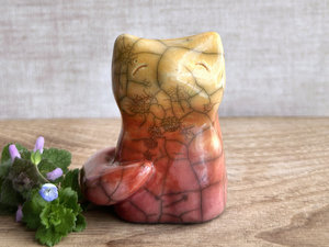 Raku ceramic sitting fox glazed in a gradient from soft bordeaux red to orange to golden yellow (bottom to top). It has a kind face, and its tail is curled around its body on its right side. There are tiny dark grey spiderweb patterns in the glaze, in addition to the normal raku crackles, from where raindrops fell on it.