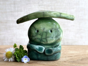 Raku ceramic sculpture glazed in forest green to spring green (bottom to top) with a kind, but slightly intense, face, and a spring-green leaf on its head. It also has two cute little arms.