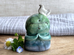 a shinto jizo nature spirit guardian raku ceramic sculpture glazed in a bottom-to-top gradient of blue to forest green to spring green. they have a happy, smiley face, little praying hands, and a sweet white bird on their head.