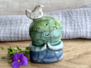 rweb patterns in the glaze, in addition to the normal raku crackles, from where raindrops fell on it.a raku ceramic jizo nature spirit guardian sculpture glazed in a bottom-to-top gradient of blue to forest green to spring green. they have a happy, smiley face, little praying hands, and a sweet white bird on their head.