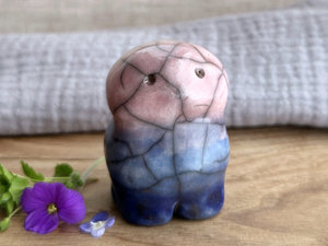 a raku ceramic creature. it has four stubby legs, a widish lower half, and a round head with a small nose and two round eyes. it tapers up a little and eventually ends in four small antlers. on the way, it has a very sweet face with a cute little nose. it's glaze in a bottom-to-top gradient of dark blue to blue to a pale pinky red. it is a friend.