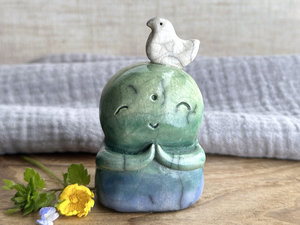 a raku ceramic jizo nature spirit guardian sculpture glazed in a bottom-to-top gradient of blue to forest green to spring green. they have a happy, smiley face, little praying hands, and a sweet white bird on their head.
