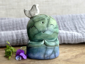 a raku ceramic jizo nature spirit guardian sculpture glazed in a bottom-to-top gradient of blue to forest green to spring green. they have a happy, smiley face, little praying hands, and a sweet white bird on their head.