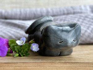 Little raku ceramic cat spirit kami sculpture glazed in grey. It has a kind, gentle, sleepy face, and its tail is curled over its back on its right.