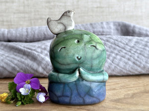 a raku ceramic jizo nature spirit guardian sculpture glazed in a bottom-to-top gradient of blue to forest green to spring green. they have a happy, smiley face, little praying hands, and a sweet white bird on their head.