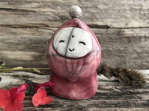 Raku ceramic gnome figure with a smiling face and a small white ceramic bauble on the top of its head. Its body is glazed in a soft Bordeaux red colour, and its face is glazed white. 