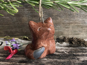 Raku ceramic kitsune fox tree ornament. It is glazed in a deep rust-brown colour, the tip of its tail is natural, unglazed black, and it has a kind, gentle face.