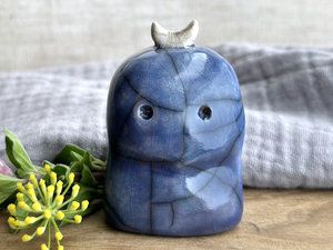 A kodama nature and forest spirit kami ceramic raku sculpture glazed in blue with faint hints of red. It has a somewhat concerned face, and a white crescent moon on its head.
