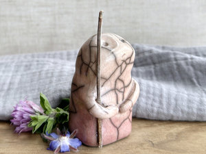 raku ceramic nature spirit warrior sculpture. it is glazed in a bottom-to-top gradient of pale soft red to pale peach and carries a brown stick in one of its arms. it has a sad and worried but determined look on its gentle face.