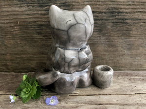 Raku ceramic sitting fox glazed in soft gradient from darker to lighter warm grey. He has a kind face, the impression of two little arms across his front like a self hug, and looks like he's sitting cross-legged.