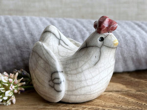 a white raku ceramic chicken with grey eyes, a pale orange beak, and a bordeaux red comb on the top of her head. she looks very sweet.