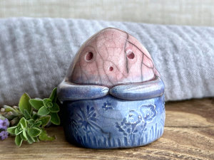 a vaguely dome-shaped nature spirit ceramic sculpture. it is glazed in a top-to-bottom gradient of soft reddish pink to blue and has plant and flower carvings on its body. it also has a gentle face, and two little arms across its front.