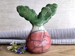 a very smiley and happy radish spirit raku ceramic sculpture. it has a round body glazed in a soft radish red colour. the top of its body fades to white, and it has two green leaves on the top of its head/body.