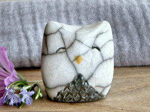 Raku ceramic little owl or owlet kami nature spirit statue with sort of pointed ears. It’s glazed in white and grey with gentle, smiling eyes and a little golden yellow beak.