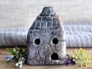 Little raku ceramic cottage glazed in muted shades of mauve, indigo, violet, purple, and brown. Its roof is carved to look like slates. It has a carved door and windows and lots of plant and flower carvings on its wall. It looks somewhat concerned.