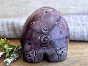 round and vaguely egg-shaped nature spirit sculpture. it is glazed in a top-to-bottom gradient of purple to violet and has spiral carvings on its body. it also has a gentle, happy face.
