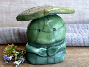 Raku ceramic sculpture glazed in a bottom-to-top gradient of forest green to spring green with a kind, but worried, face, and a golden yellow and green leaf on their head. They have two cute little arms.