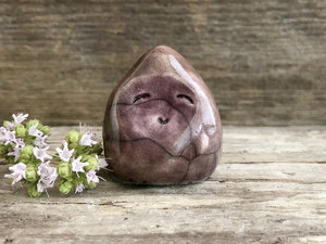 A small, almost (but not quite) teardrop-shaped raki ceramic earth spirit sculpture. It has a gentle face and is glazed in soft purple and warm brown colours.