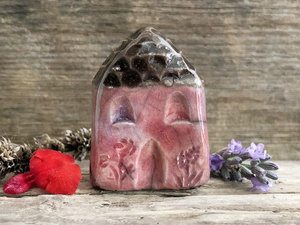 Tiny raku ceramic cottage glazed in soft deep red, violet, and mauve. Its roof is glazed in grey-brown and carved to look like slates. It has a carved door and windows and lots of plant and flower carvings too.