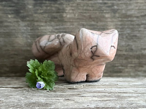 Raku ceramic kitsune fox sculpture glazed in a soft warm peach colour. It has a kind, gentle face, and a tail curled over its back. There are tiny dark grey spiderweb patterns in the glaze, in addition to the normal raku crackles, from where raindrops fell on it.
