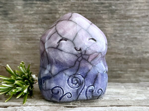 a nature spirit sculpture shaped a bit like a wide hourglass that's a little pointy at the top. it is glazed in a bottom-to-top gradient of indigo-violet to soft mauve and has spiral carvings on its body. it also has a gentle, happy face.