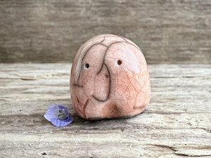 a small and stout nature spirit raku sculpture glazed in a peach colour. it has a cute little face, and a little plant carving on its right side.