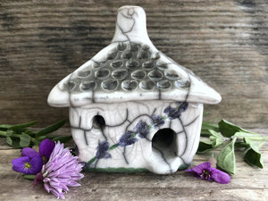 Raku ceramic kurinuki cottage glazed in white with green and lavender flower patterns on the walls. The roof has a chimney and is glazed in white and grey to look like slate roof tiles. It has a cute little doorway and lots of windows.