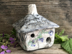 Raku ceramic kurinuki cottage glazed in white with green, purple, and violet flower patterns on the walls. The roof has a chimney and is glazed in grey, white, and black to look like slate. It has a cute little doorway and lots of windows.
