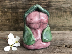 Raku ceramic sculpture of a girl with a green hood and green petal arms. Her face and body are glazed in a muted deep pink, and she has a gentle smile.