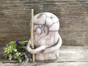 raku ceramic nature spirit warrior sculpture. it is glazed in very pale, soft mauve colour and carries a brown stick in one of its arms. it has a sad and worried but determined look on its gentle face.