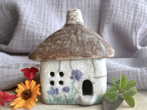 a rustic raku ceramic kurinuki cottage glazed in white with green plant carvings and purple, violet, and indigo flower carvings on the walls. the roof has a chimney and is glazed in brown with hints of white and carved to look like thatch. the cottage has a cute little doorway and lots of windows.