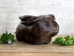 Raku ceramic sculpture of a rabbit. It is sitting with its long ears folded down its back and is glazed in dark brown. It has a very sweet, gentle face.