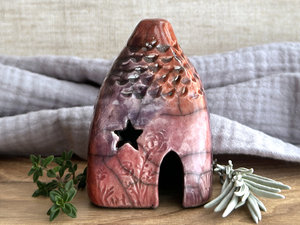 Raku ceramic kurinuki cottage glazed in bordeaux red, warm orange, mauve, and purple. It has a cute little chimney, plant and flower carvings on its walls, a cute door, and little windows, one of which is carved in a star shape. Its roof is carved to look like tiles.