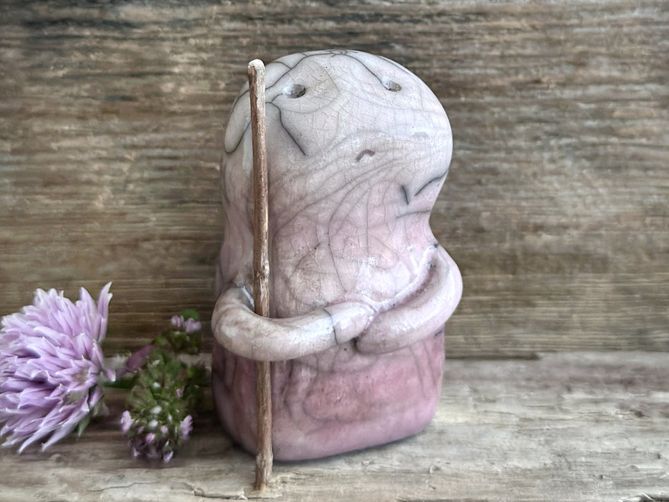 raku ceramic nature spirit warrior sculpture. it is glazed in a gradient from darker to lighter soft mauve plum (bottom to top) and carries a brown stick in one of its arms. it has a sad and worried but determined look on its gentle face.