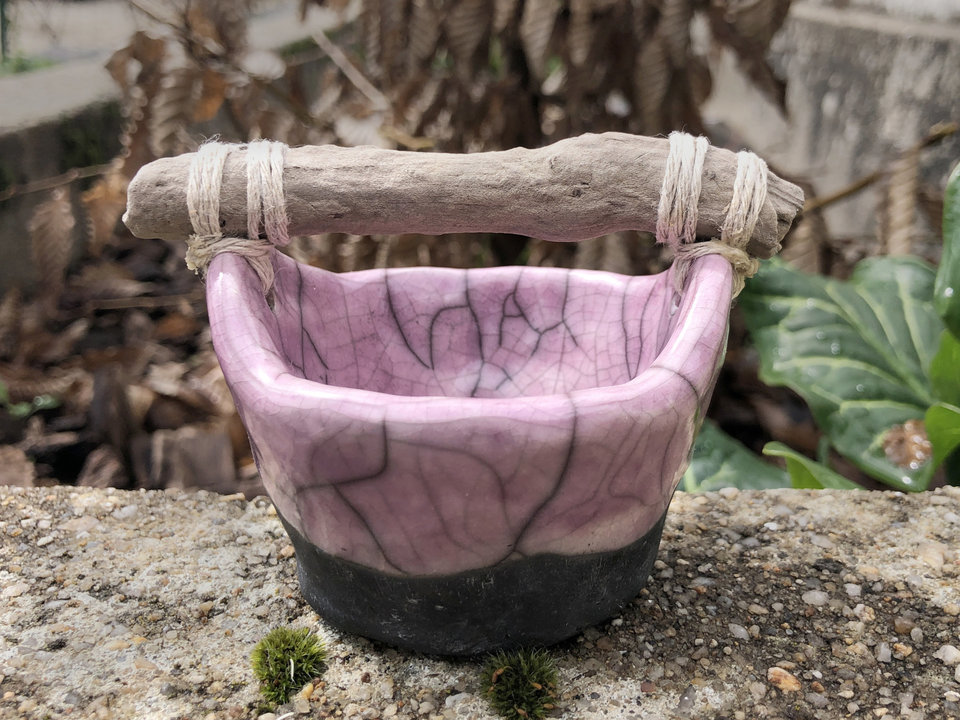Small raku ceramic bucket with a driftwood handle. The bucket is glazed a mauve pink on the inside and halfway down the outside, and the rest of the outside is left natural, unglazed black.