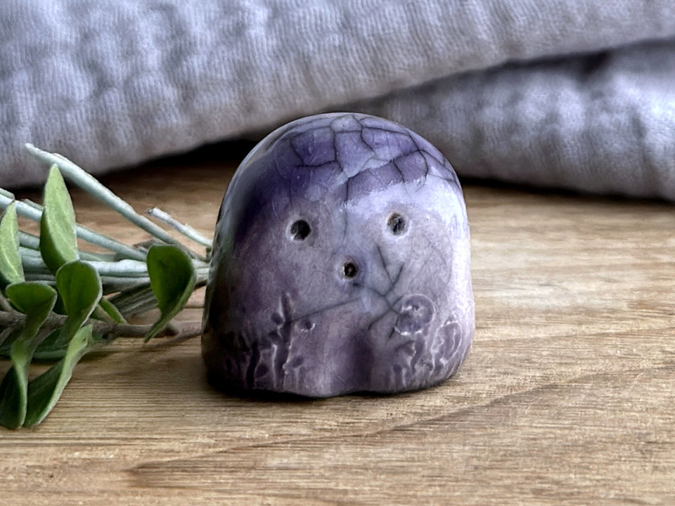 a small and stout nature spirit raku sculpture glazed in violet purple. it has a cute little face, and flower and plant carvings on its front.