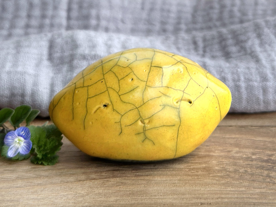 a very happy raku ceramic sculpture of a lemon. it's glazed in a golden yellow colour. it has a friendly face and lemon-skin dimpling on its body.