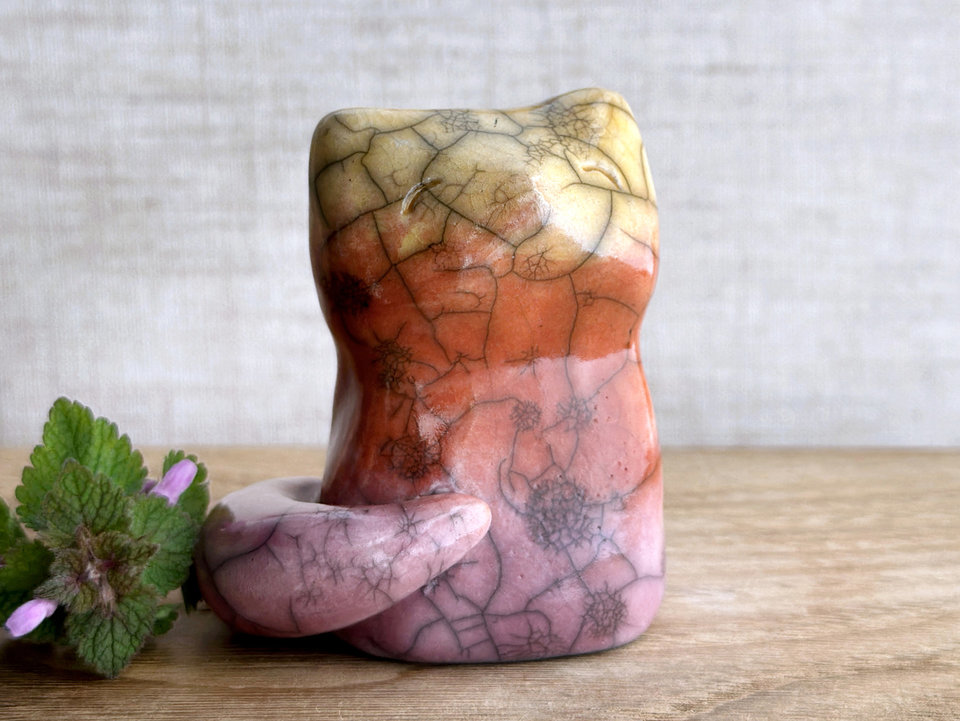 Raku ceramic sitting fox glazed in a gradient from mauve to orange to golden yellow (bottom to top). It has a kind face, and its tail is curled around its body on its right side. There are tiny dark grey spiderweb patterns in the glaze, in addition to the normal raku crackles, from where raindrops fell on it.